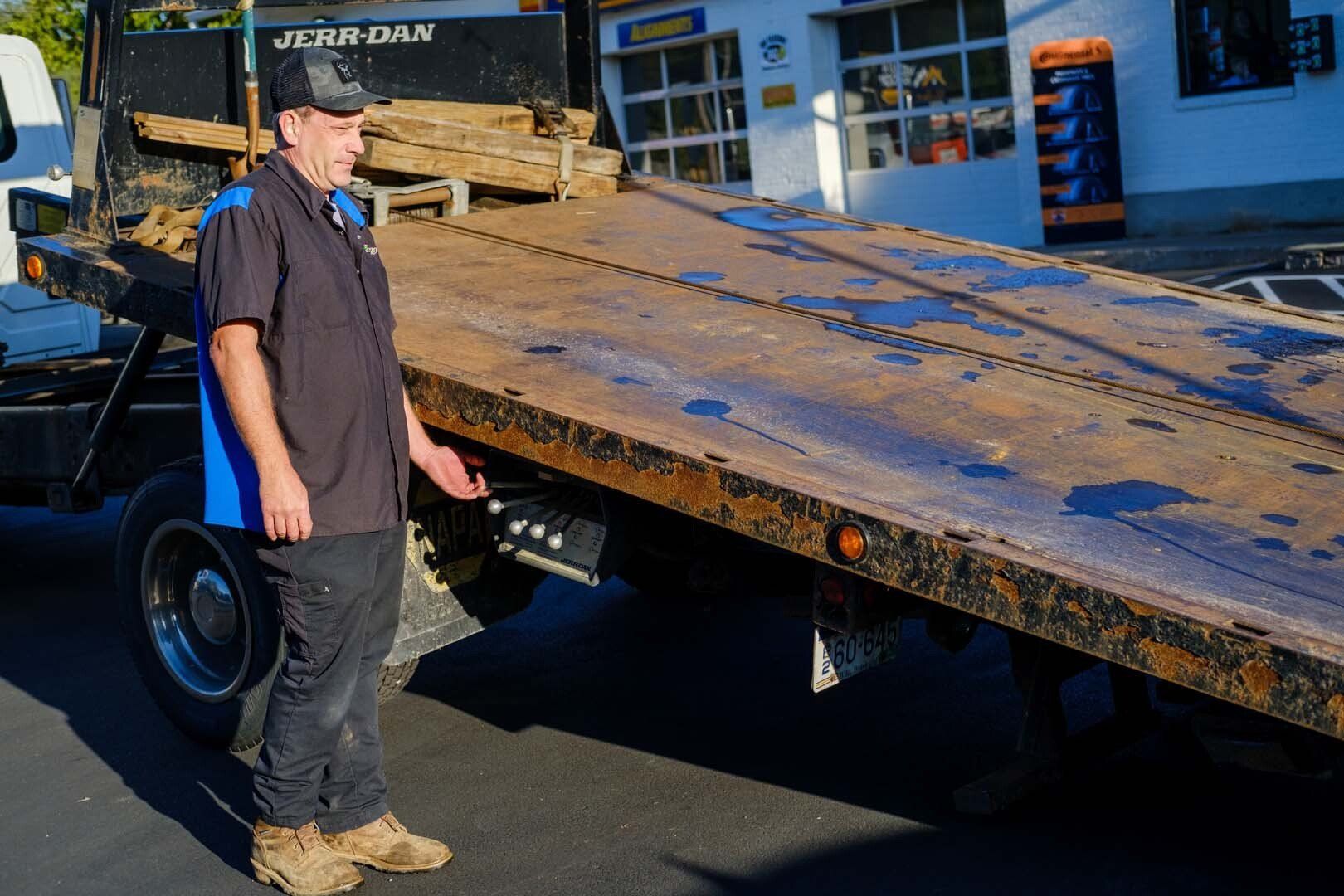 Man in uniform stands by a tow truck's flatbed. The flatbed is weathered. A shop is in the background.