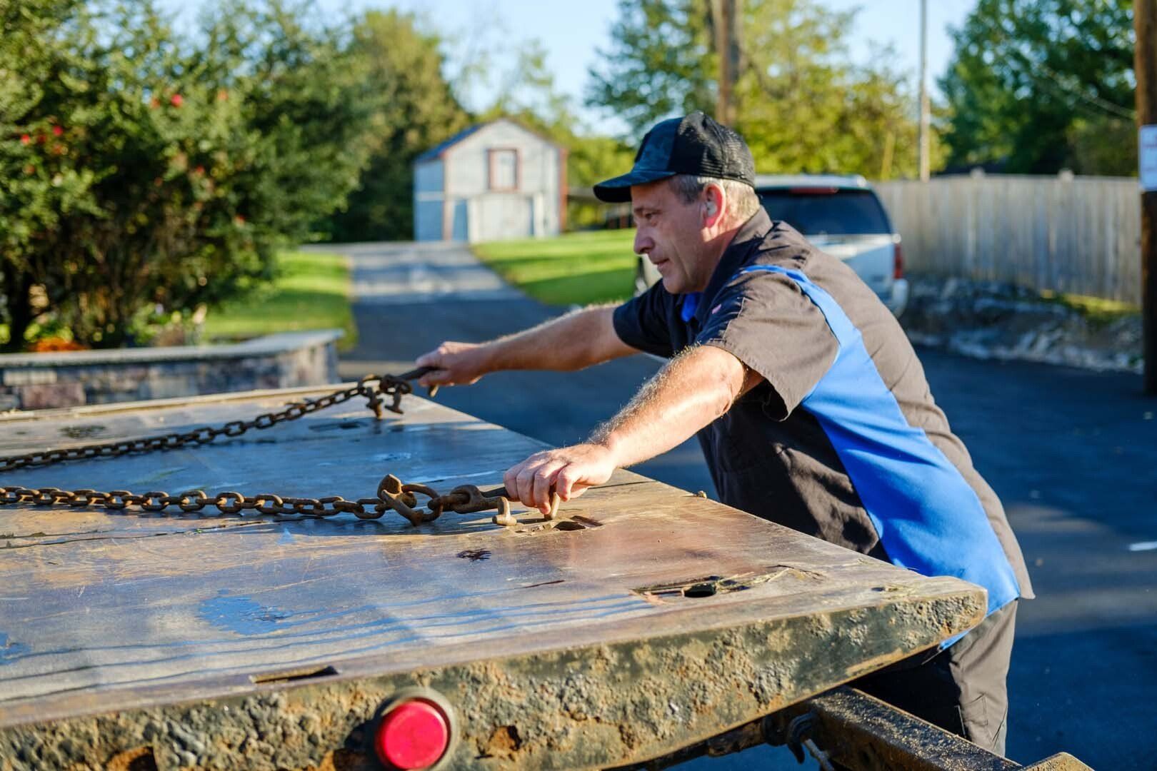 Man in a mechanic's uniform securing chains on a tow truck bed outdoors.