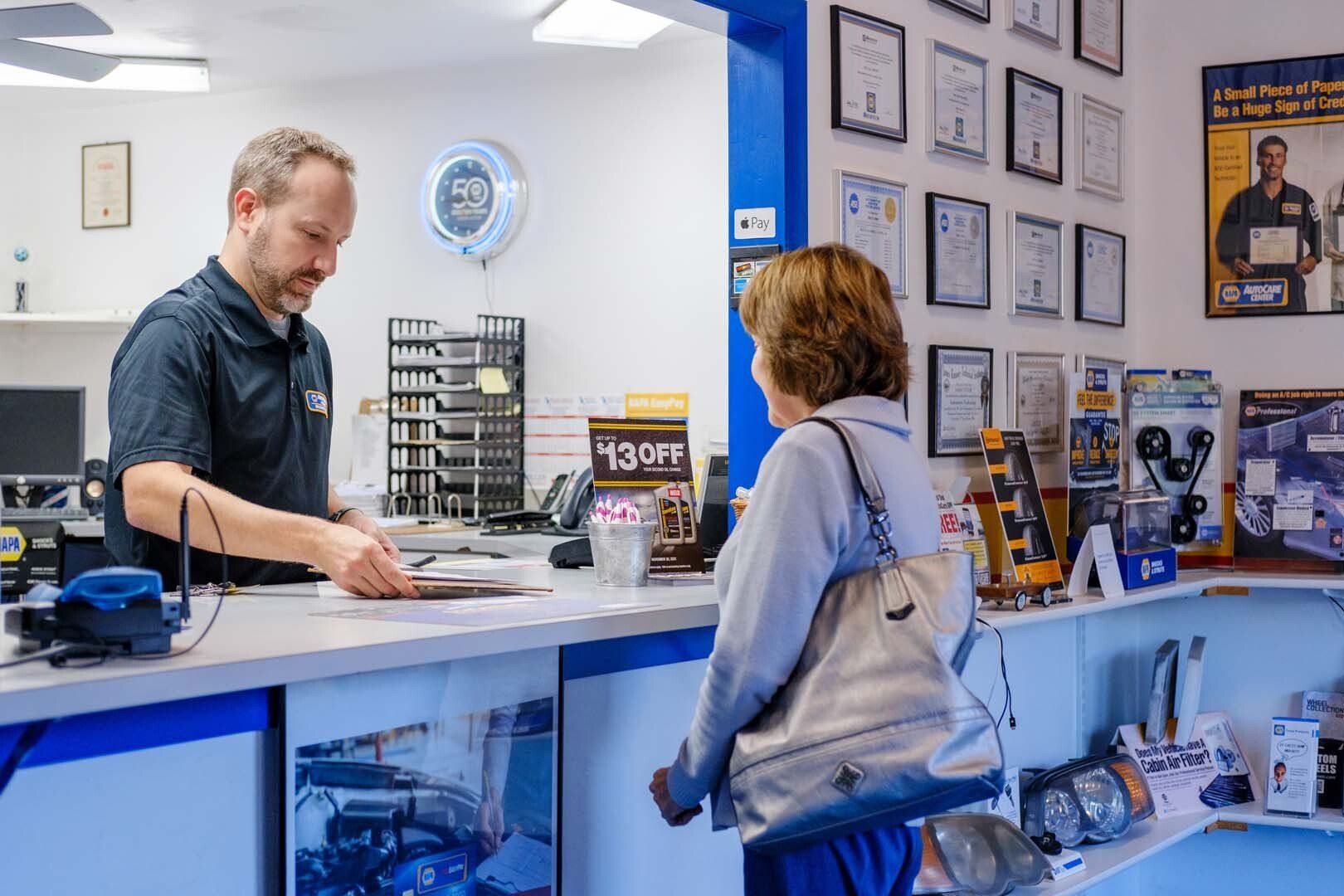 Man assisting a customer at an auto shop counter. Papers are exchanged. Blue and white colors.
