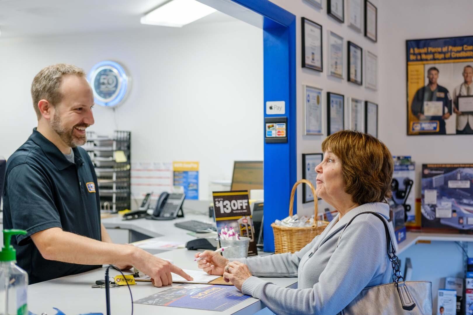 Man pointing to paperwork, smiling at a woman at an auto repair shop counter. Blue wall, awards, and supplies are in the background.