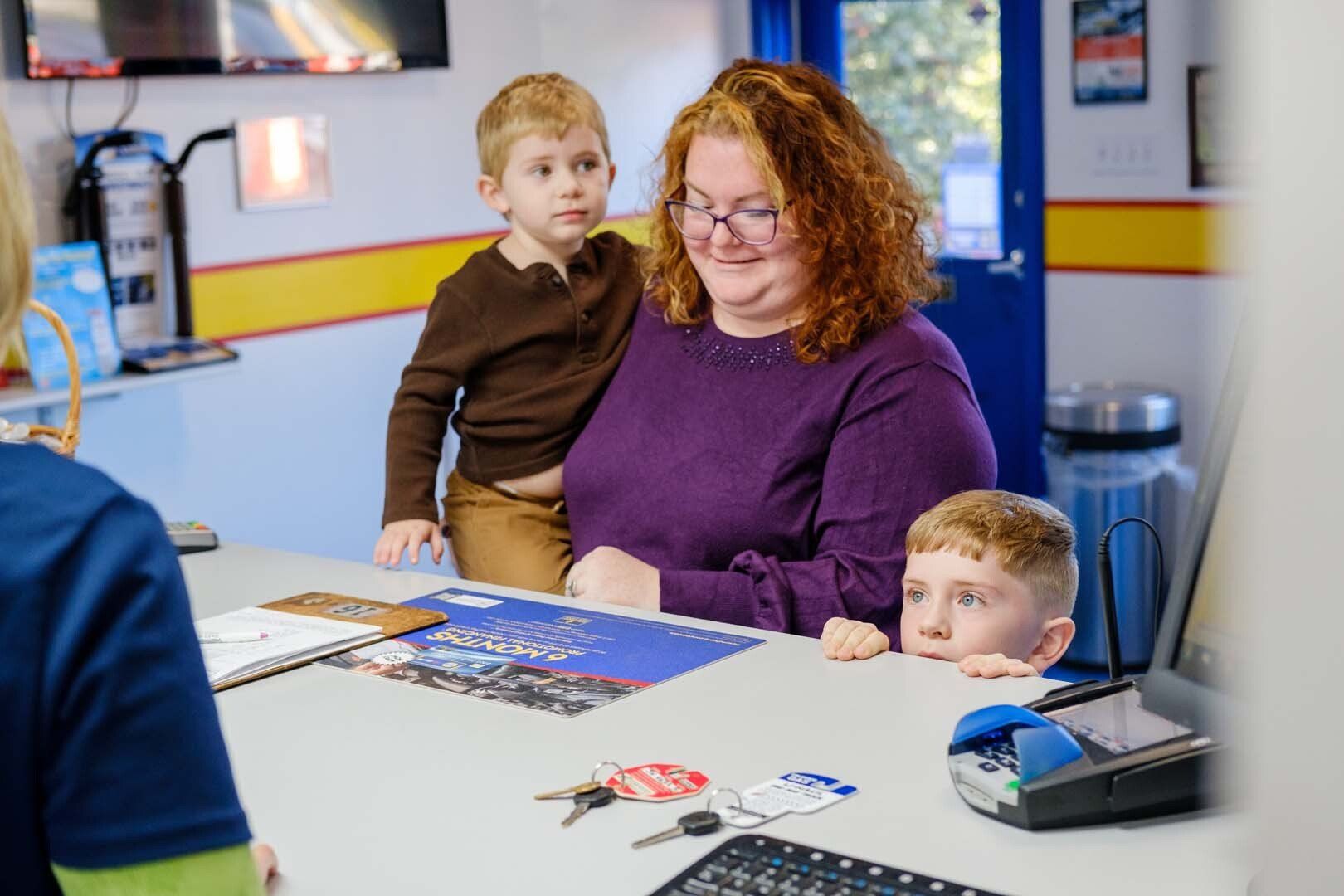 Woman with two children at a counter; one child peeking, the other standing behind her. Inside auto shop.