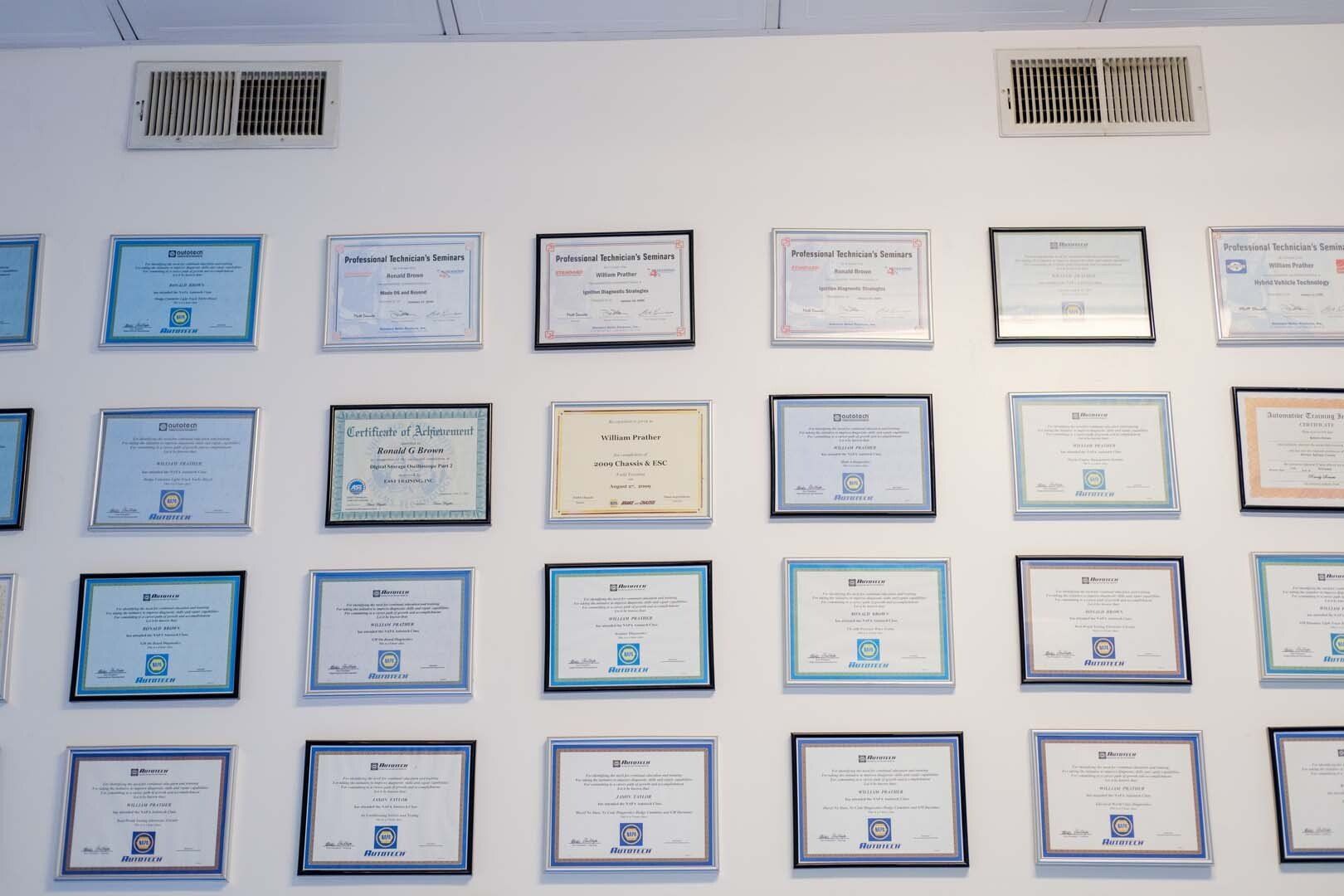 Wall display of framed certificates, varying colors, in a white-walled interior.