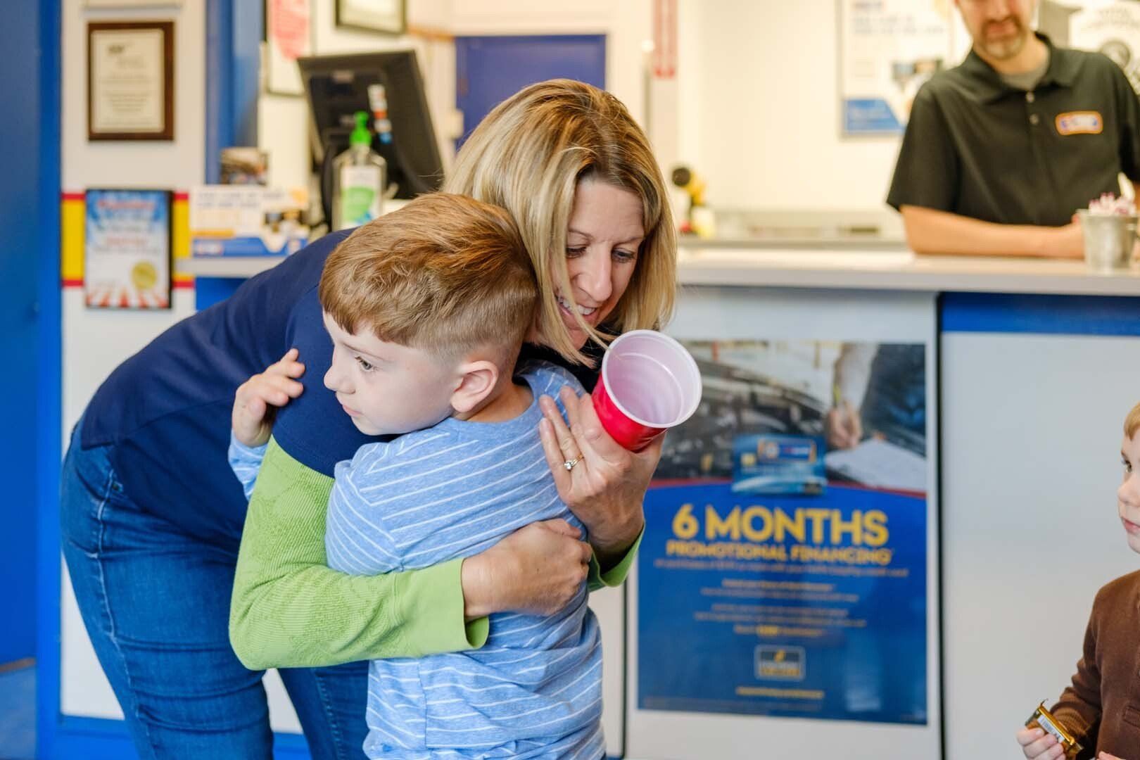 Woman hugs a child at a car repair shop counter, holding a red cup; another person stands behind the counter.