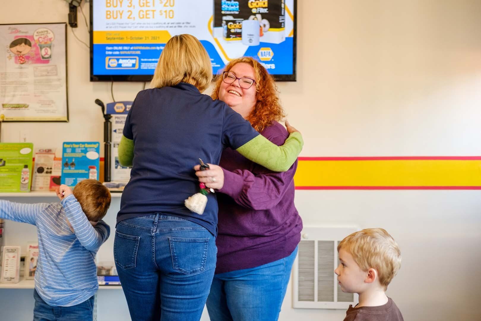 Two women embrace inside a store, with two children nearby. One woman wears jeans, the other a purple shirt.