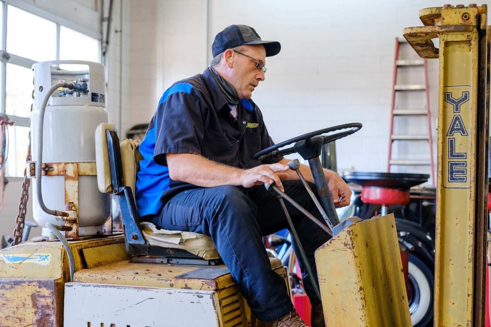 Mechanic in blue shirt and cap operating a yellow Yale forklift inside a garage.