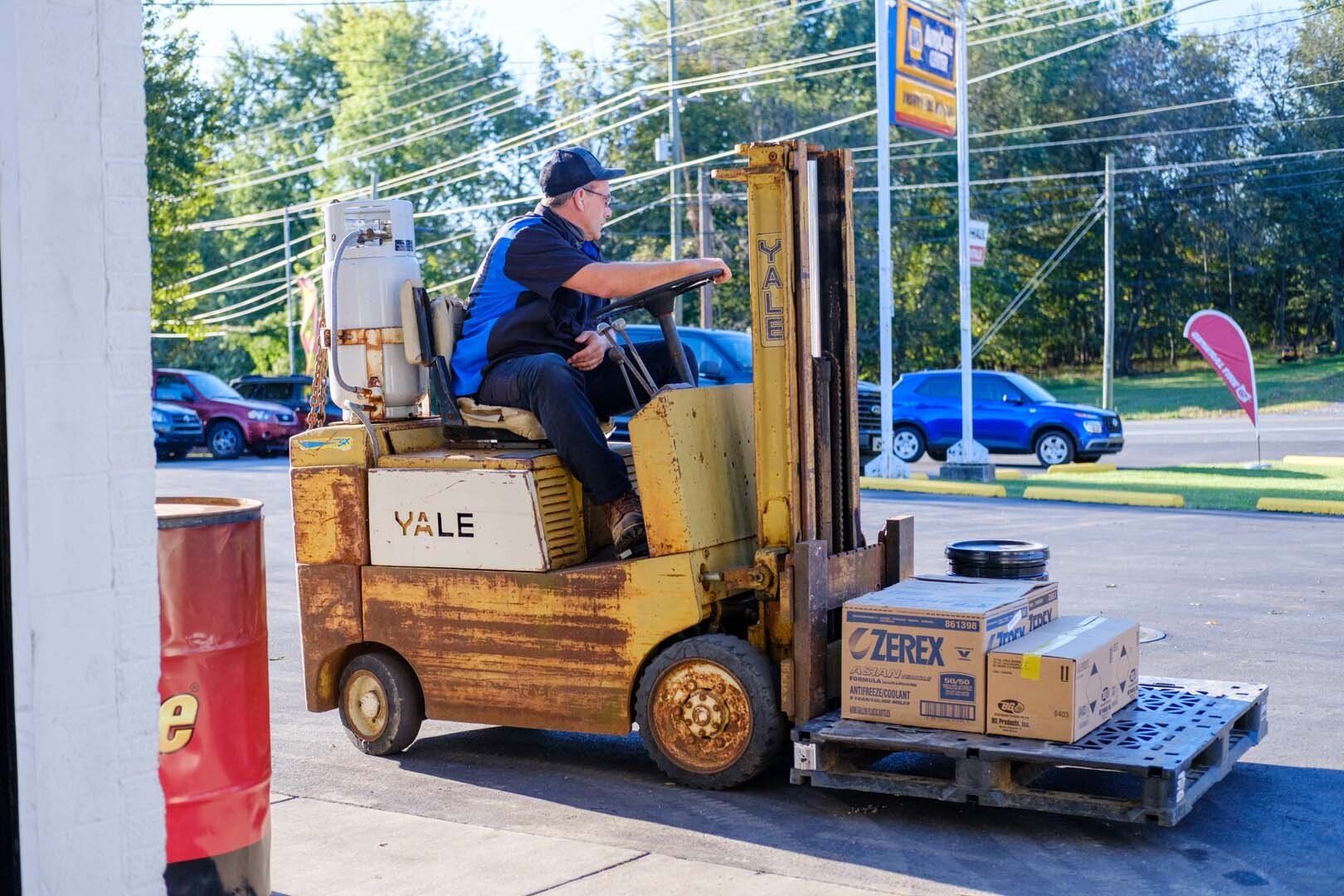 Person operating a yellow Yale forklift, carrying boxes on a pallet outside a gas station.