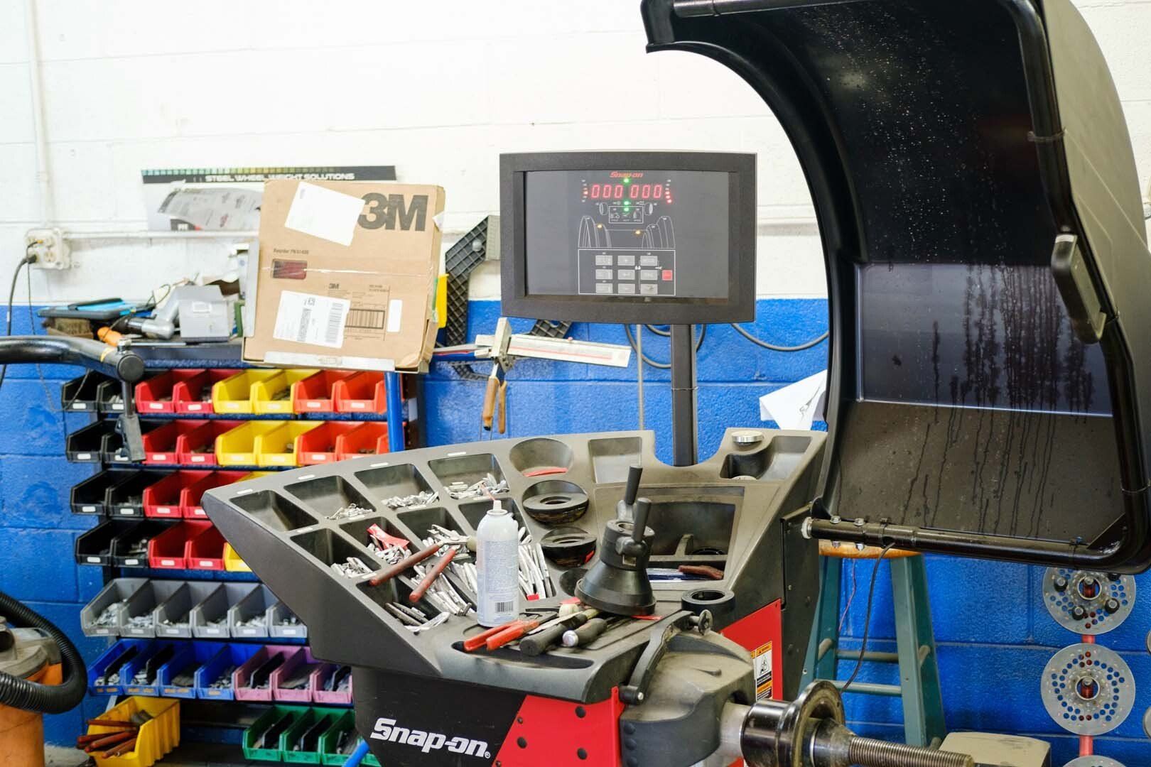 Tire balancing machine in auto shop, display showing readings, tools and parts visible, blue wall and storage shelves in background.