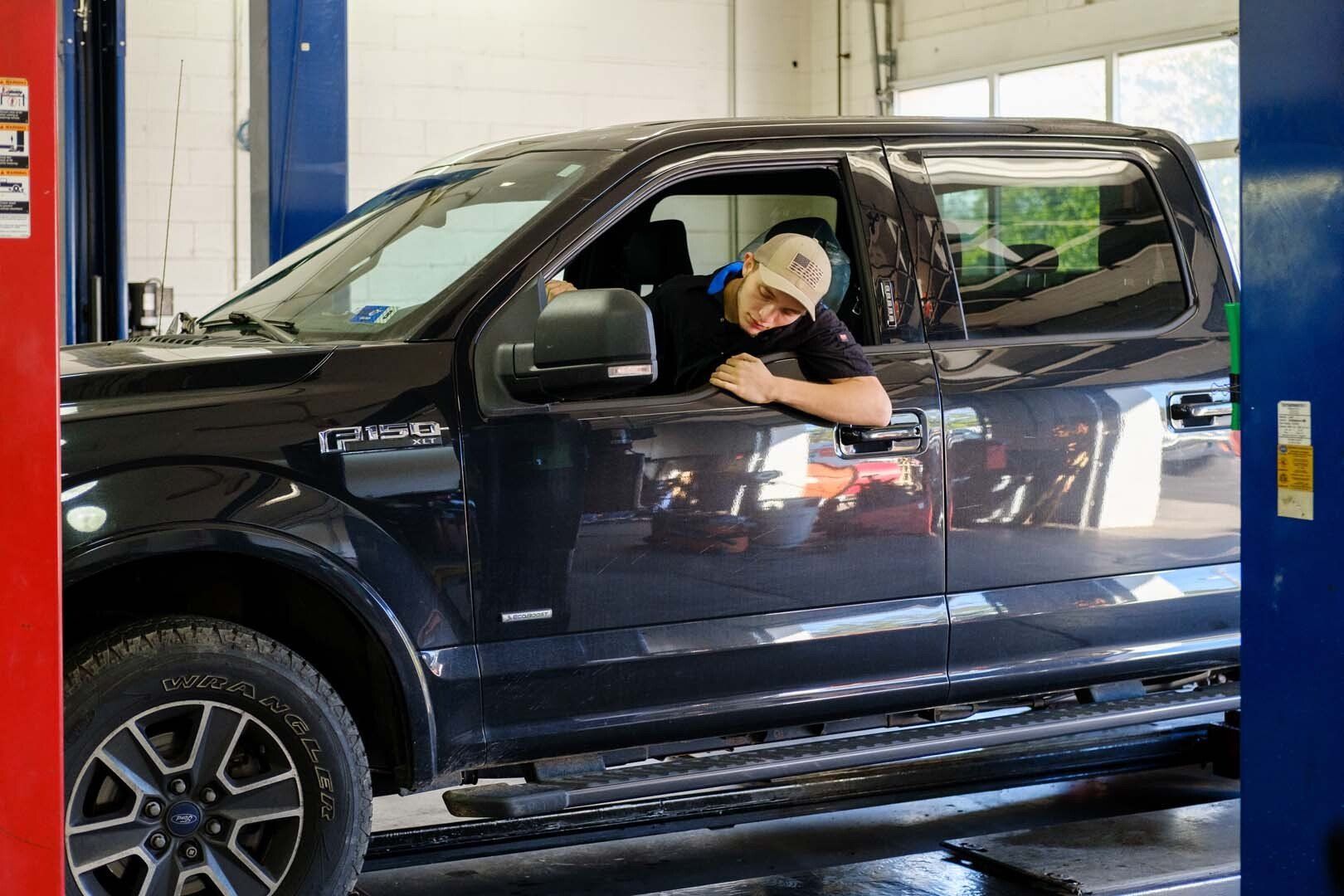 Man in a baseball cap leaning out of the window of a black truck in a garage.