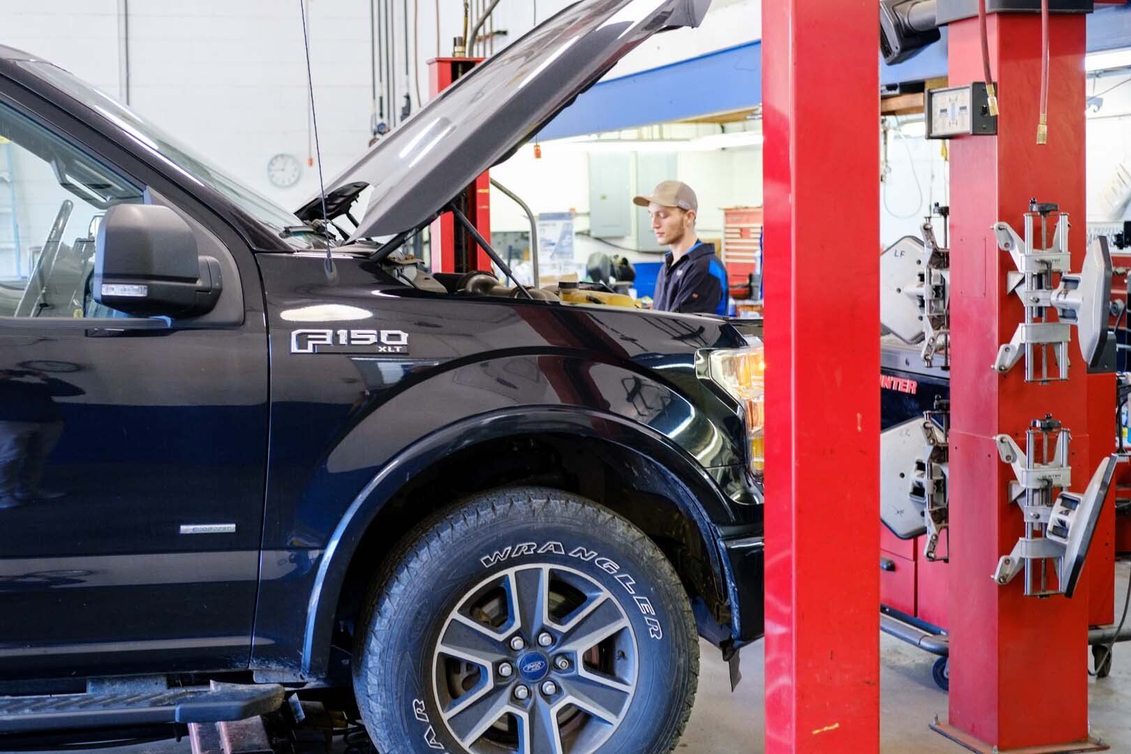 Black Ford F-150 with hood open in auto shop, mechanic in background, red support column.