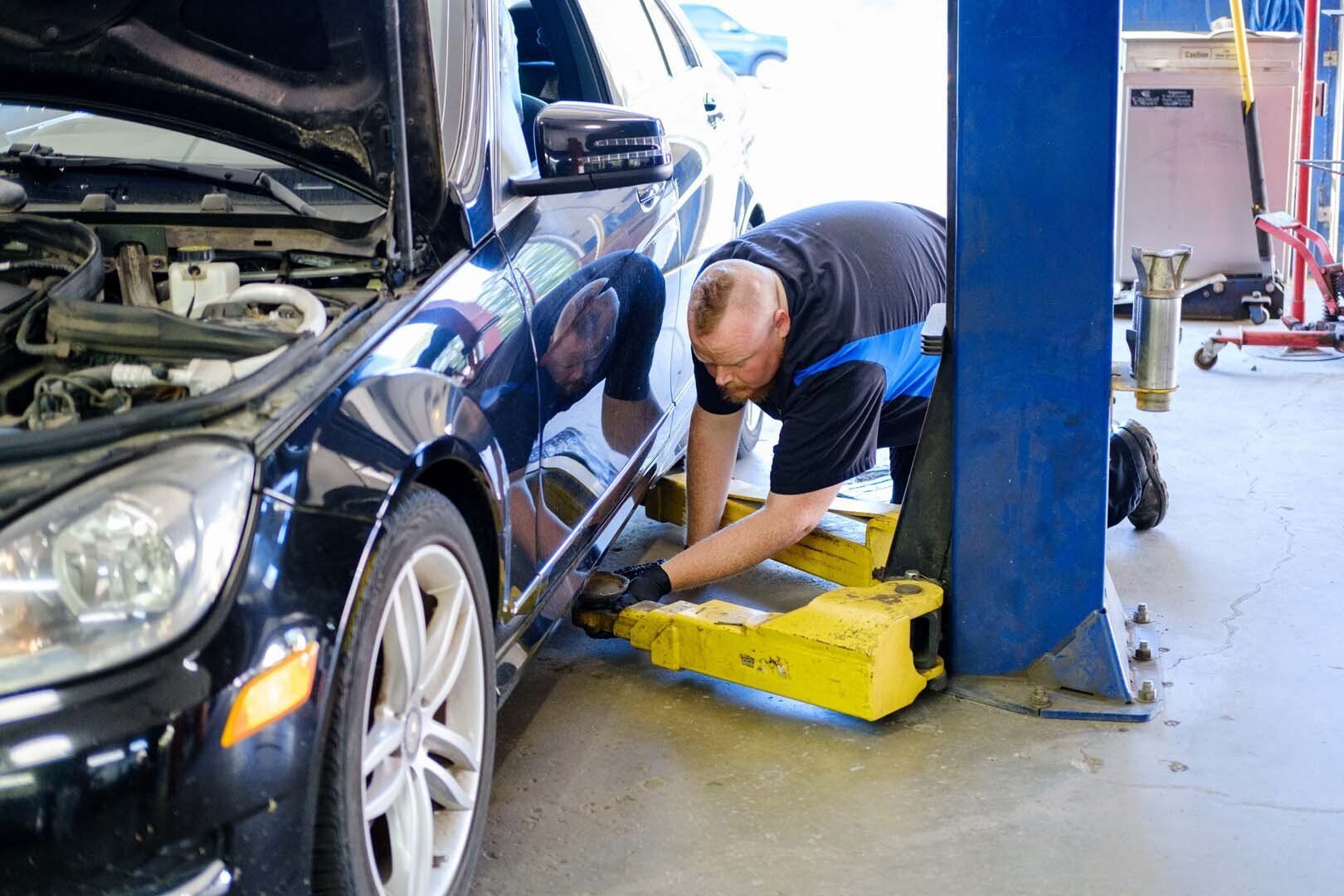 Mechanic working on a black car lifted on a yellow hoist in a garage.