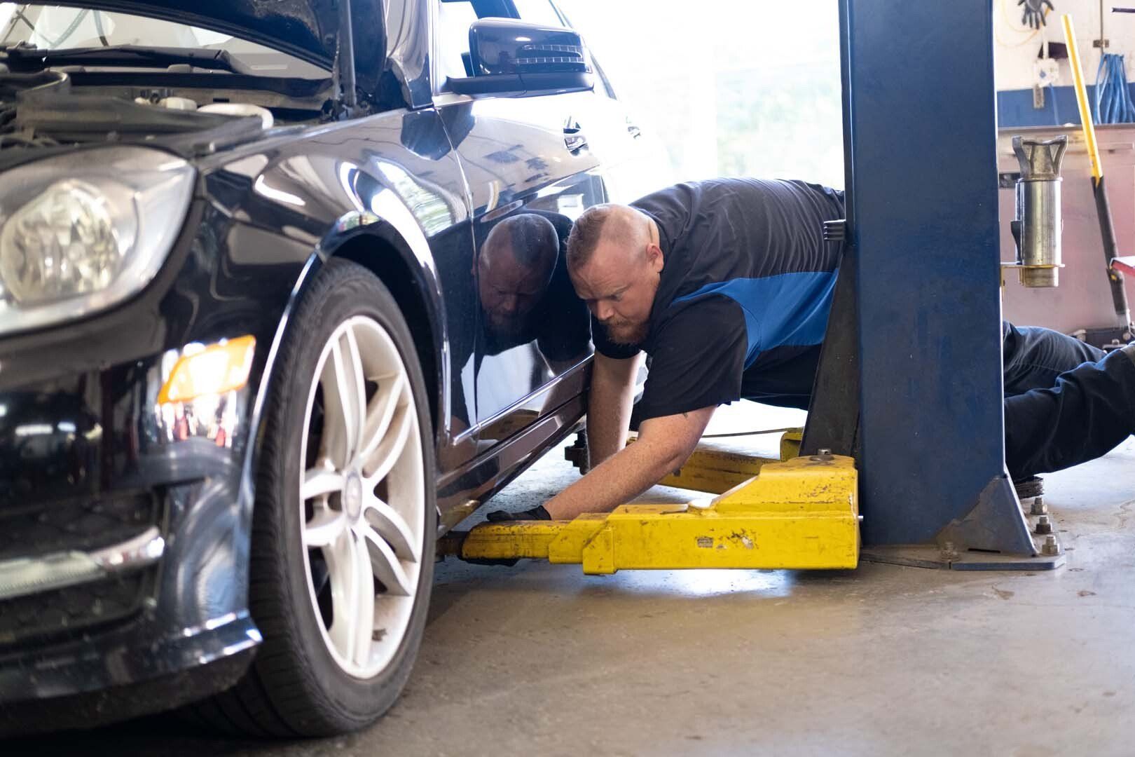 Mechanic working on a black car lifted on a yellow hoist in a garage.