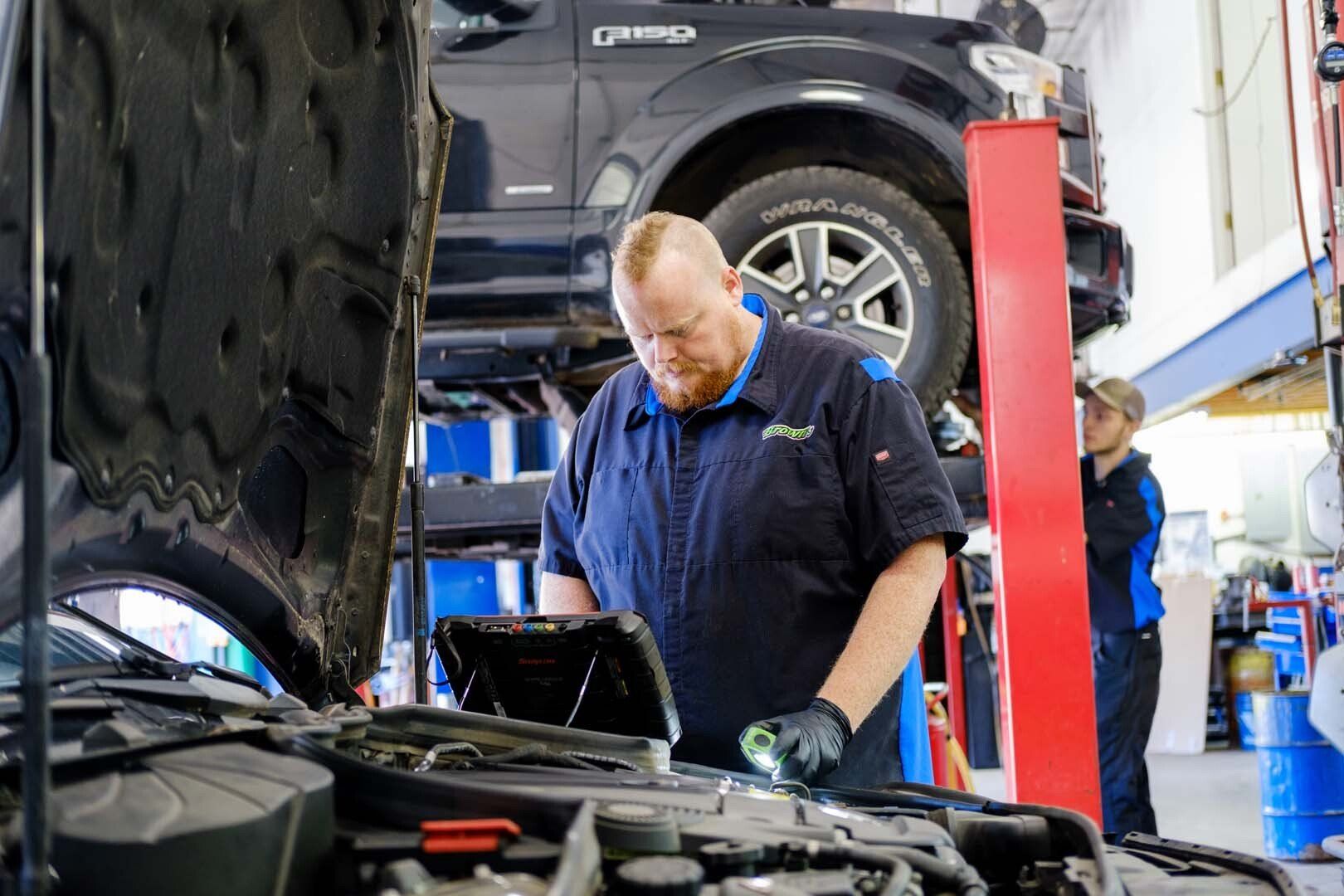 Mechanic inspecting a car engine with a flashlight, another person in the background, vehicle on a lift.