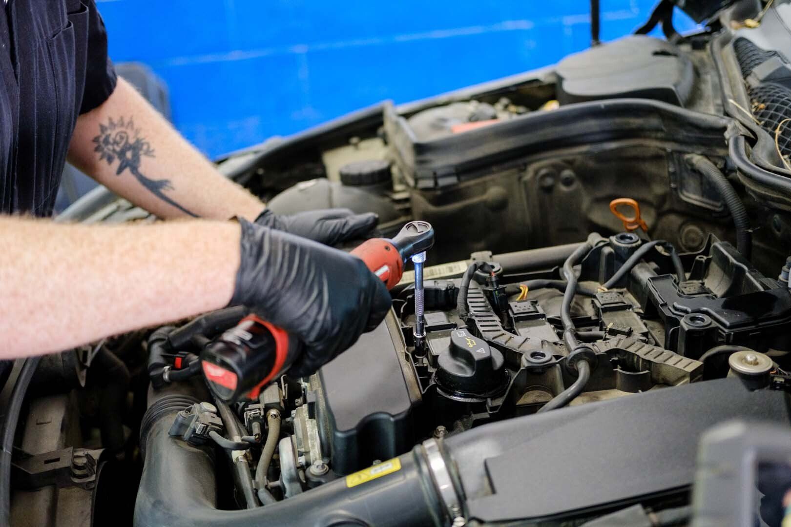Mechanic using a wrench on a car engine, wearing gloves, in a garage with a blue wall.