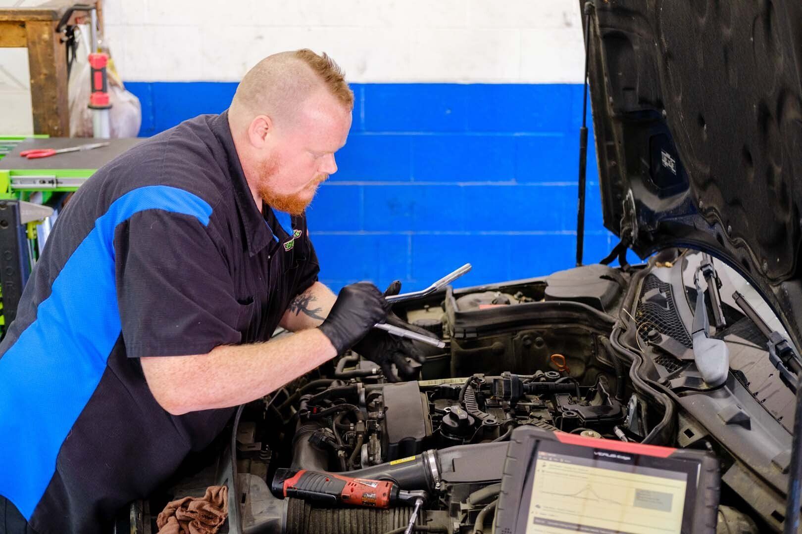 Mechanic working on car engine in a shop, using tools and a diagnostic tablet. Blue and black work uniform.