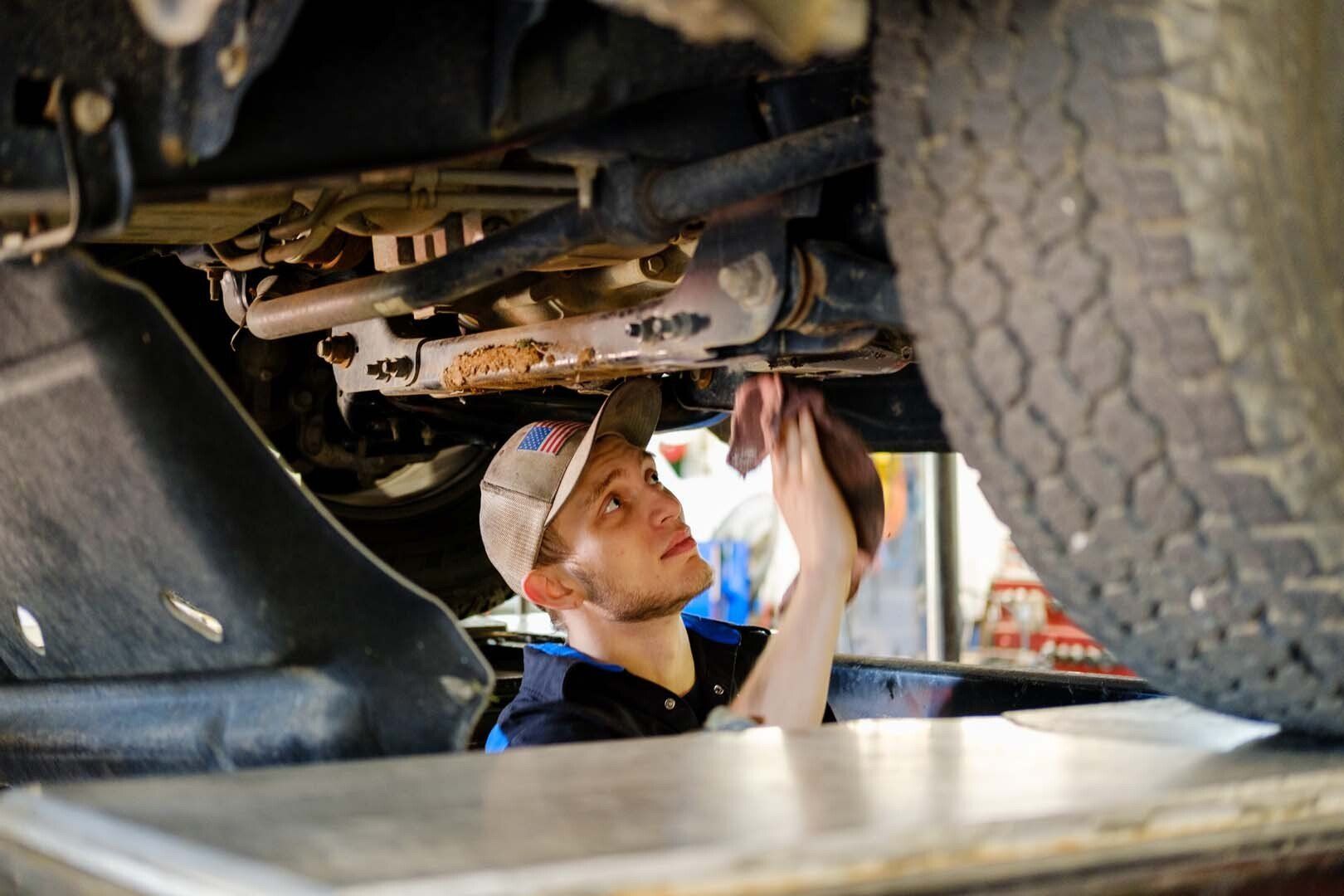 Mechanic working under a vehicle; using a rag. He's wearing a cap and is in a garage setting.
