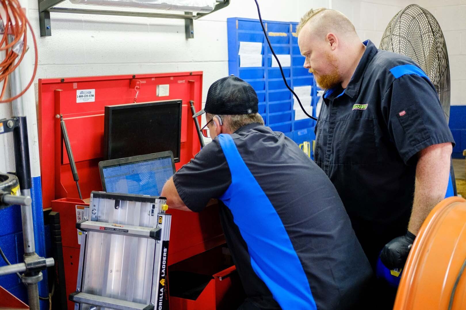 Two mechanics looking at a computer in a red cabinet in a garage. One is wearing a black hat.