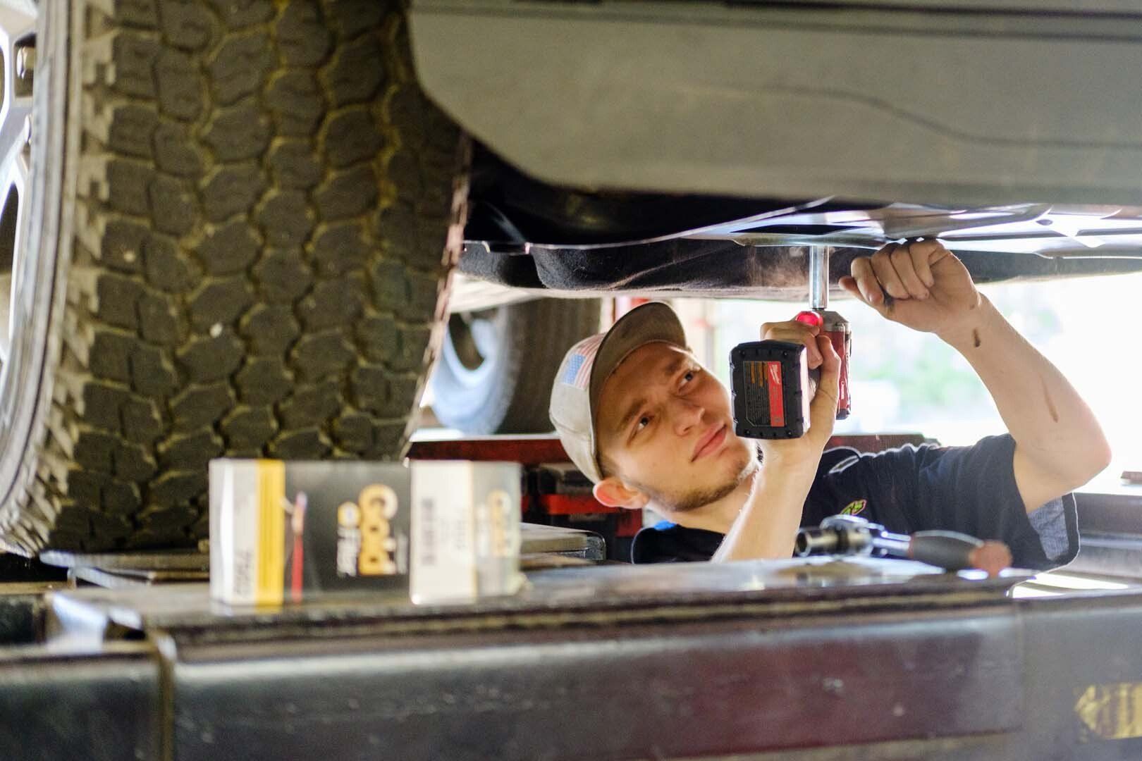 Mechanic working under a car, using a tool. He wears a baseball cap. A tire is visible on the left.