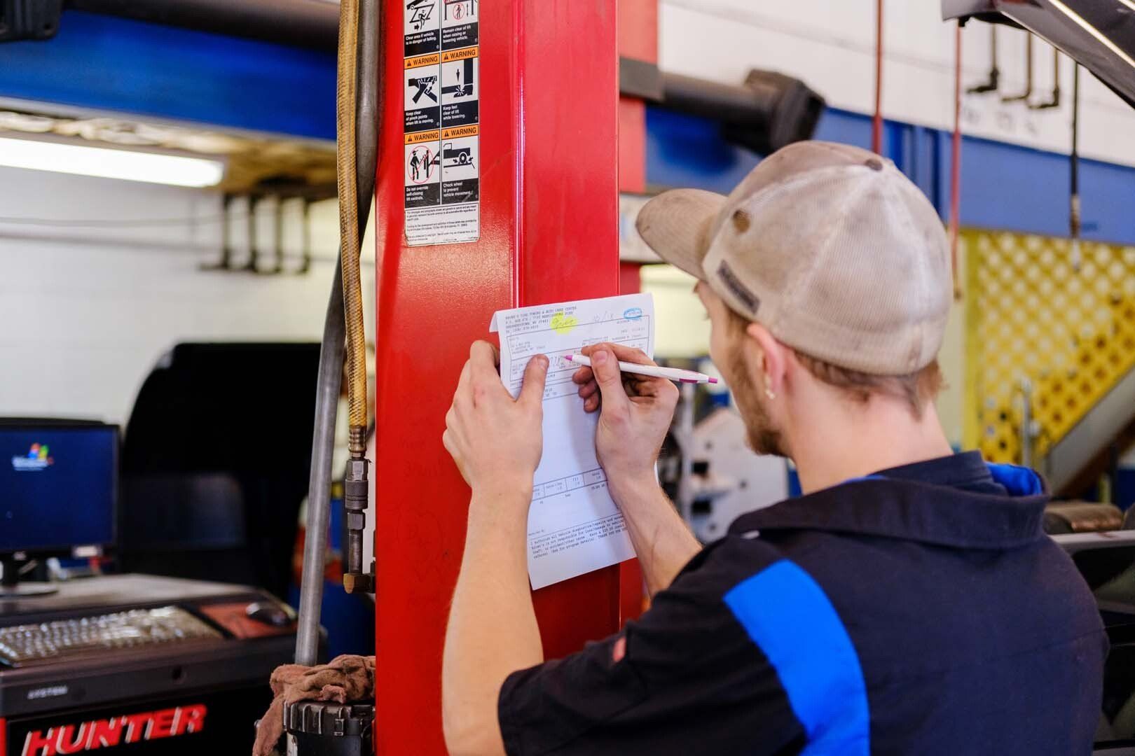 Mechanic in a tan cap and work shirt, writing on a notepad attached to a red lift post in a garage.