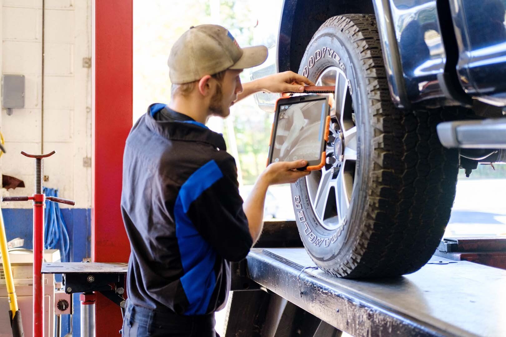 Mechanic using a tablet to inspect a car tire on a lift in a shop.
