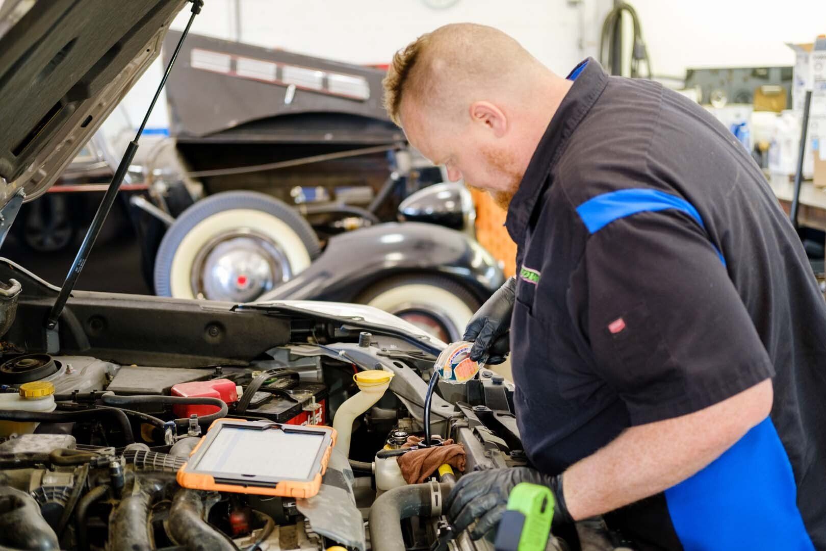 Mechanic examines engine with tablet and flashlight. Car is in a garage.