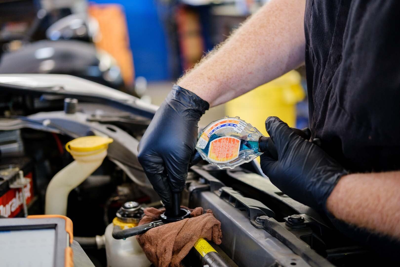 Mechanic with black gloves, working on a car engine, holding a radiator cap in an auto shop.