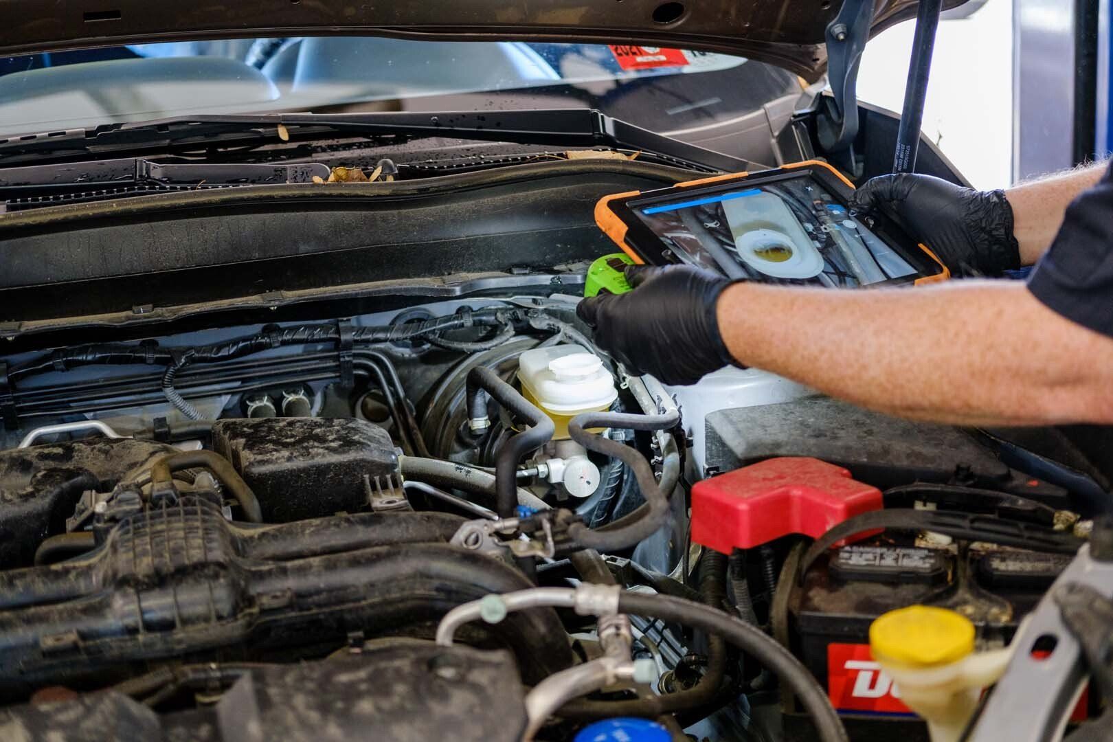 Mechanic using a tablet to diagnose a car engine under the hood. Black gloves and open hood.