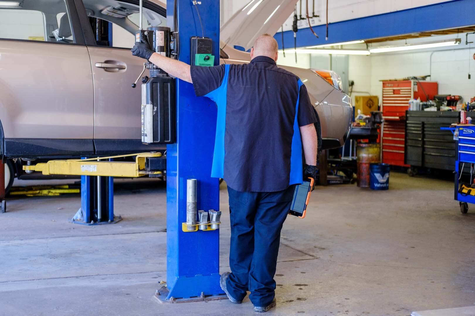Mechanic in blue and black shirt leans on a blue lift post, holding a tablet in auto shop.