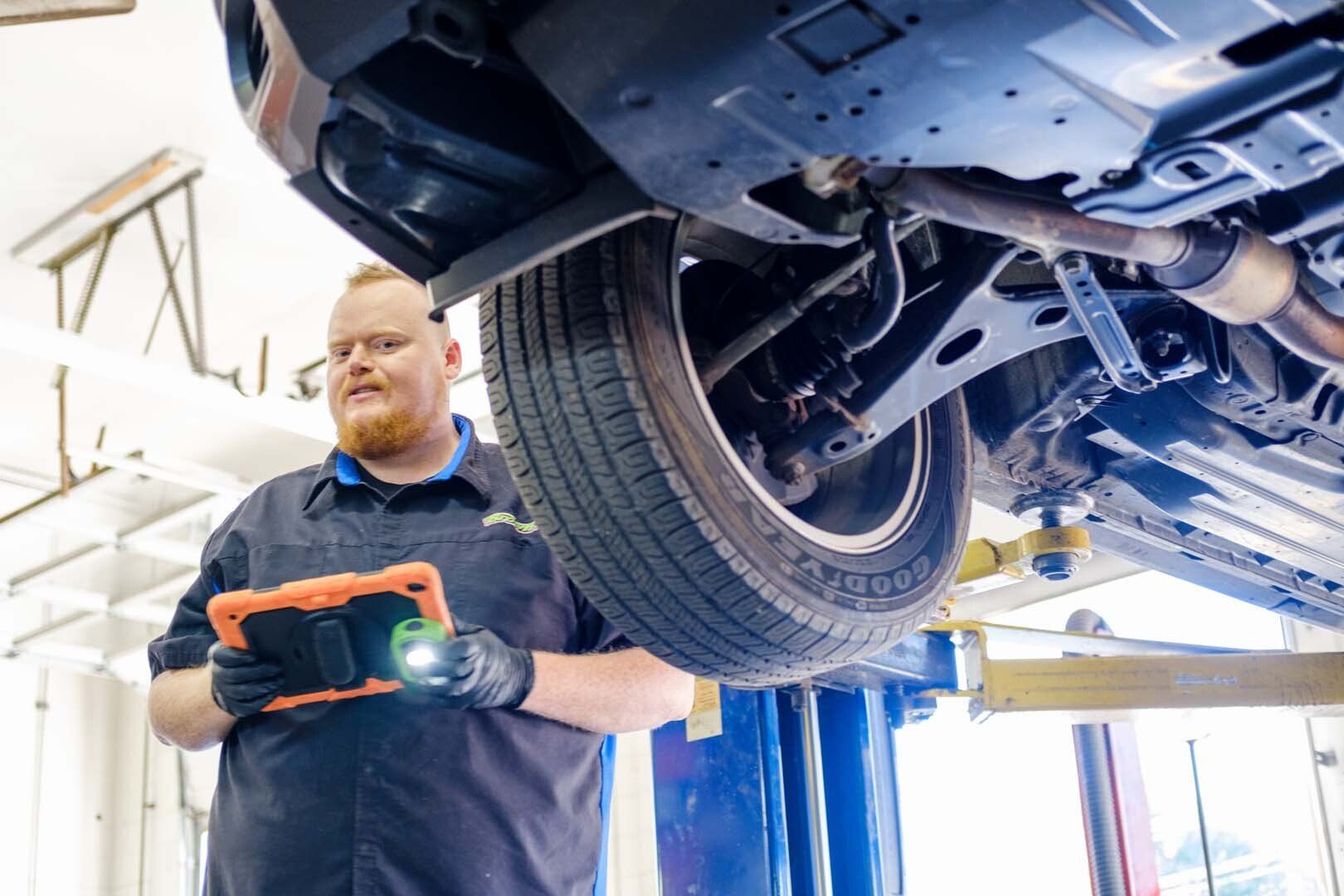 Mechanic inspecting under a car raised on a lift, using a tablet and flashlight.