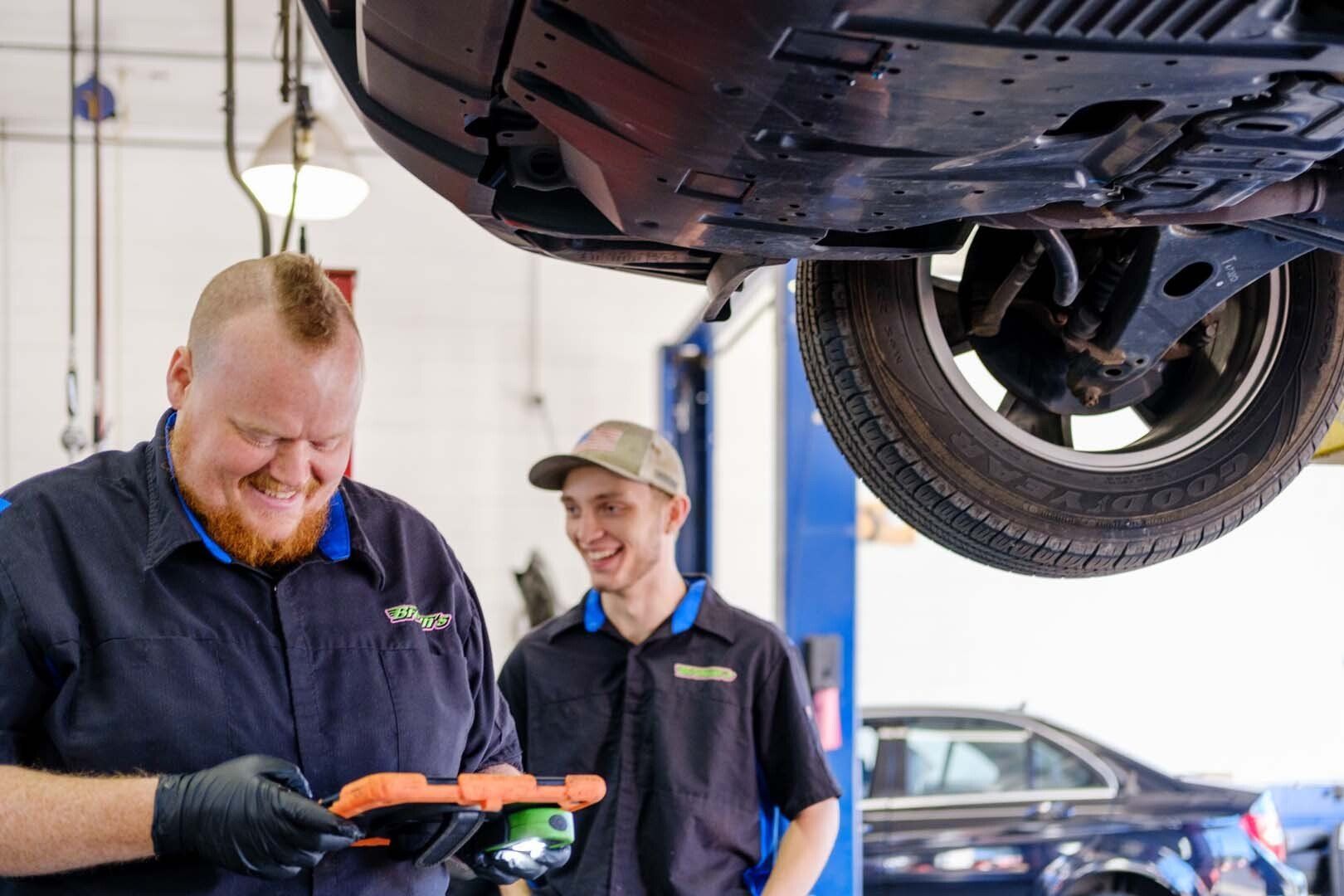 Two mechanics working under a car, one smiling at a diagnostic tool.
