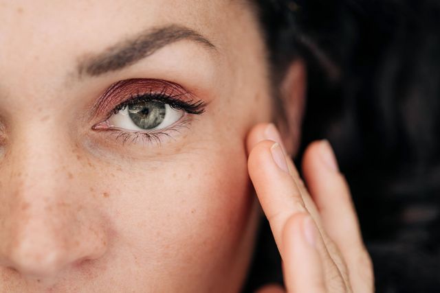 Primo piano di una donna con lentiggini e ombretto rosso, che si tocca il viso vicino all'occhio.