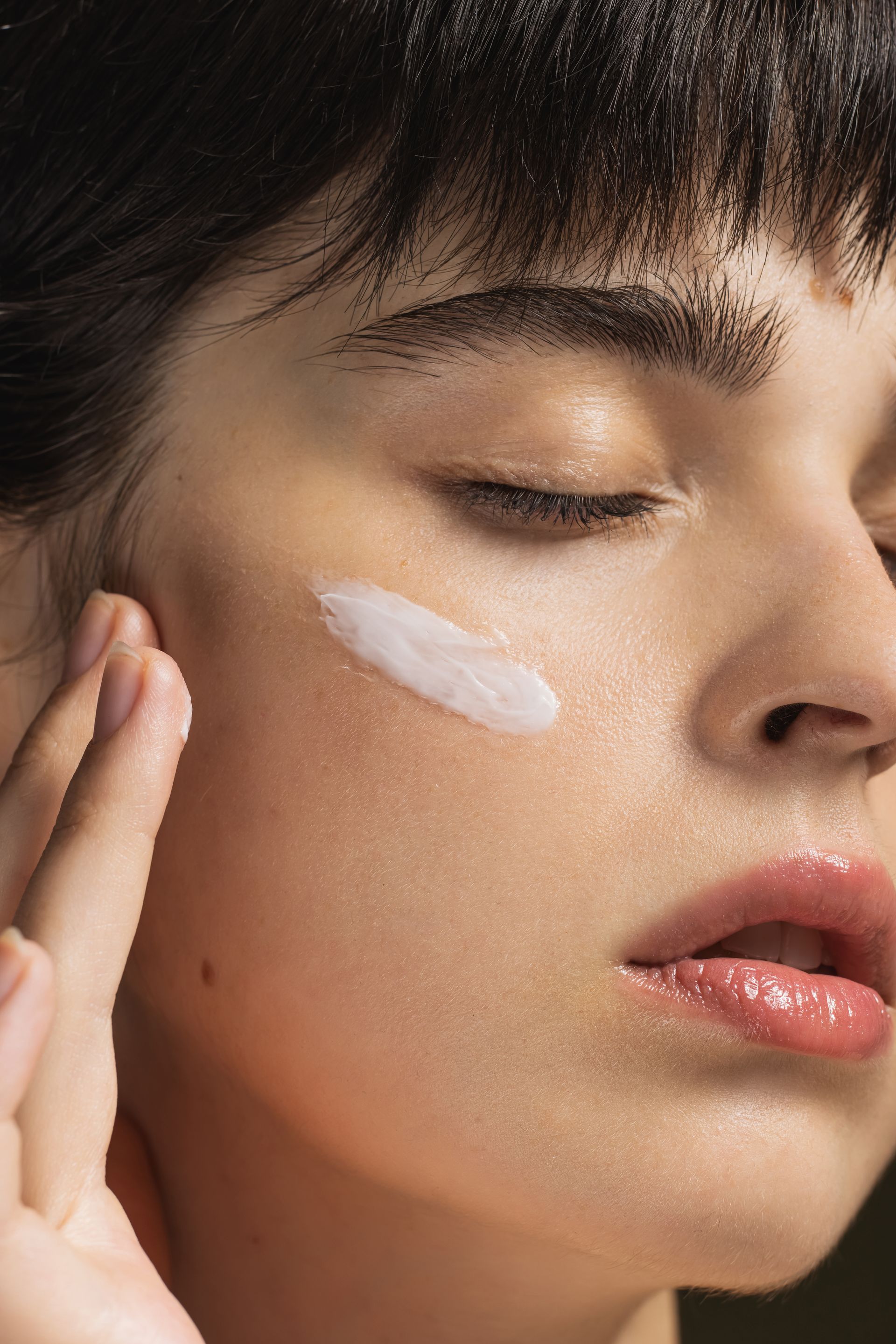 Donna che si applica la crema sulla guancia, occhi chiusi, luce soffusa.