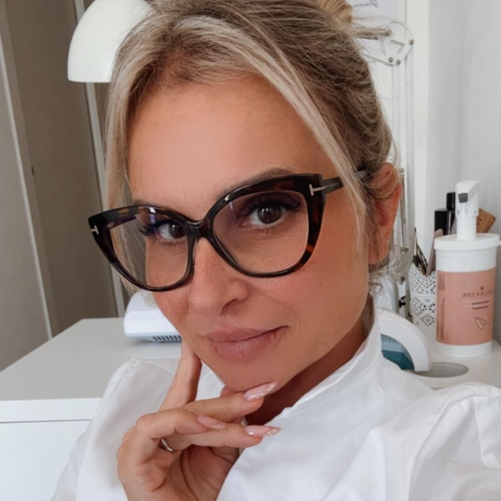 Una donna bionda con gli occhiali sorride, con la mano sul mento, in una stanza ben illuminata.