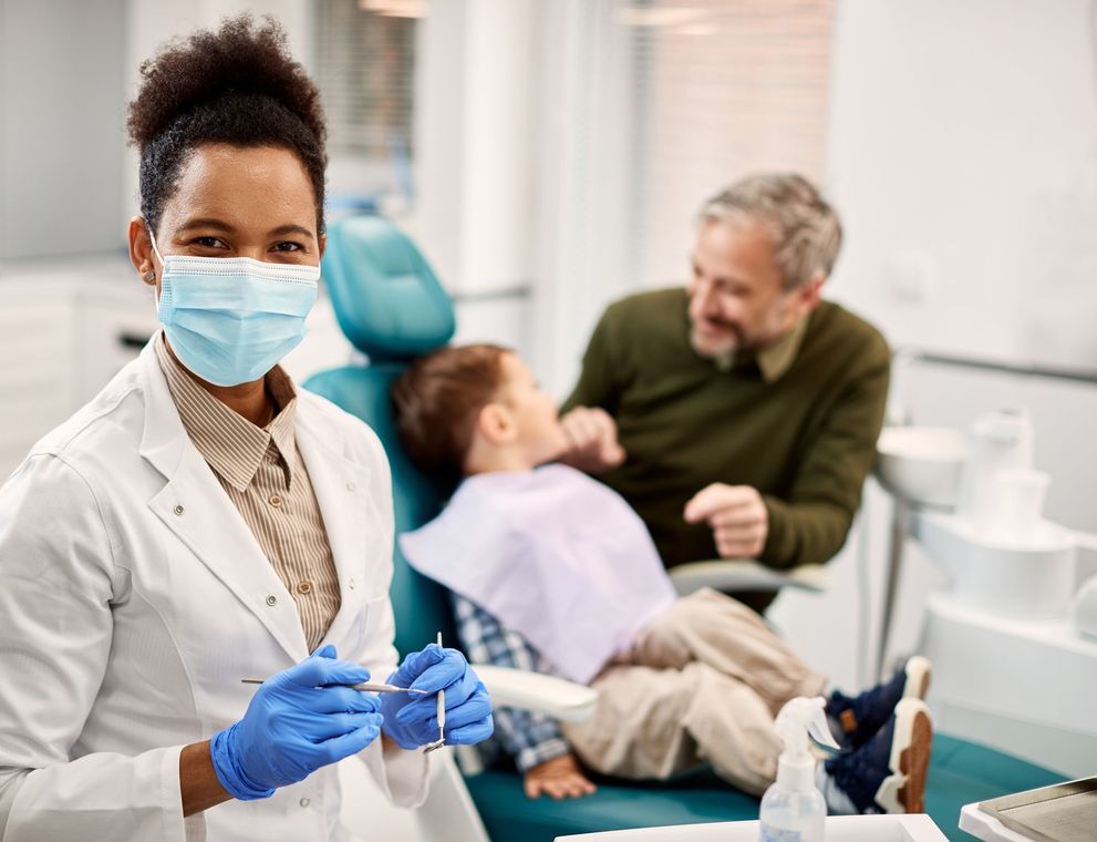 A female dentist wearing a mask and gloves is standing in front of a child in a dental chair.