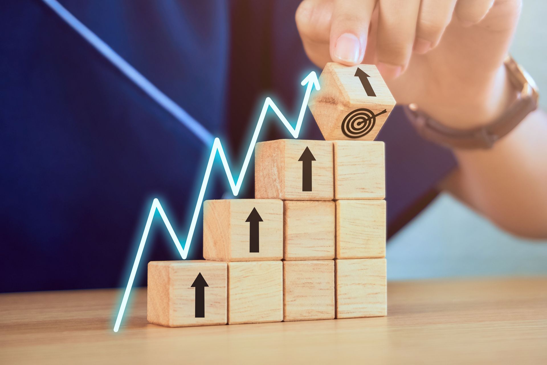 Hand placing a wooden block with a target on top of a stack of blocks with upward arrows; progress graph overlaid.