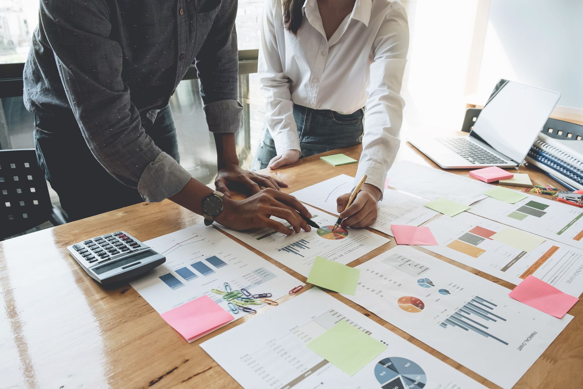 Two people working at a wooden desk, reviewing business documents, charts, and sticky notes for a project.