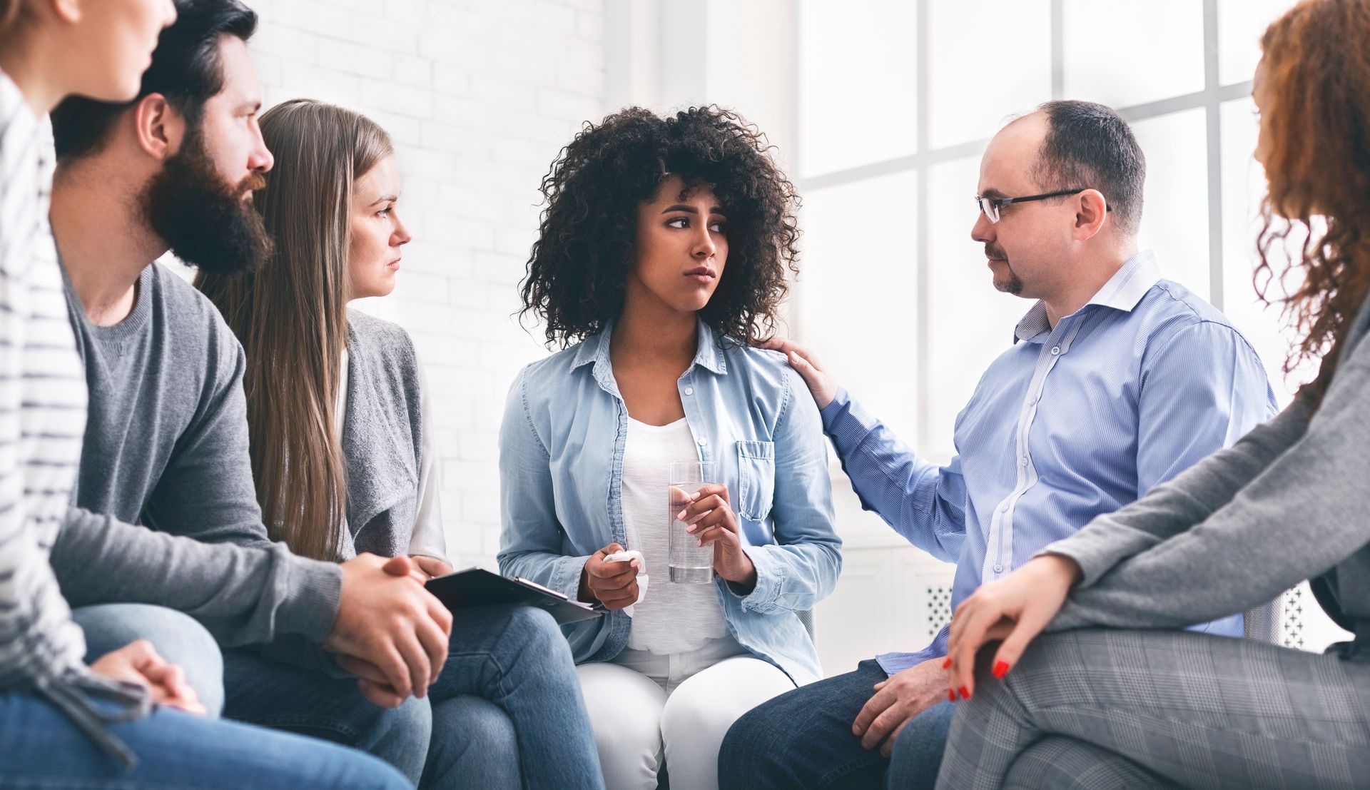 A group of people sit in a circle in a bright room for a support meeting, one person offering comfort to another.