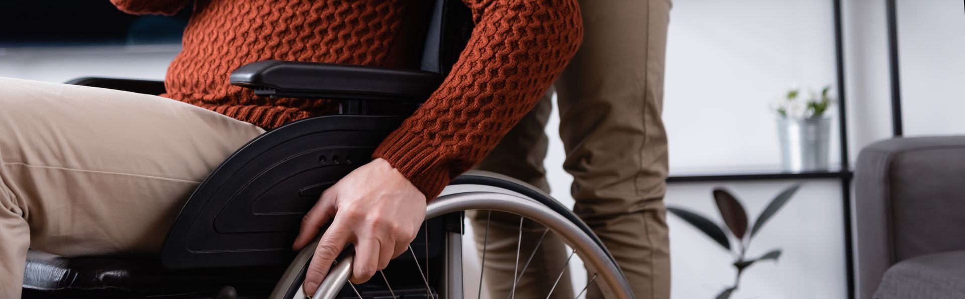 A caregiver in blue scrubs stands behind an individual in a wheelchair, looking at them in a brightly lit room.