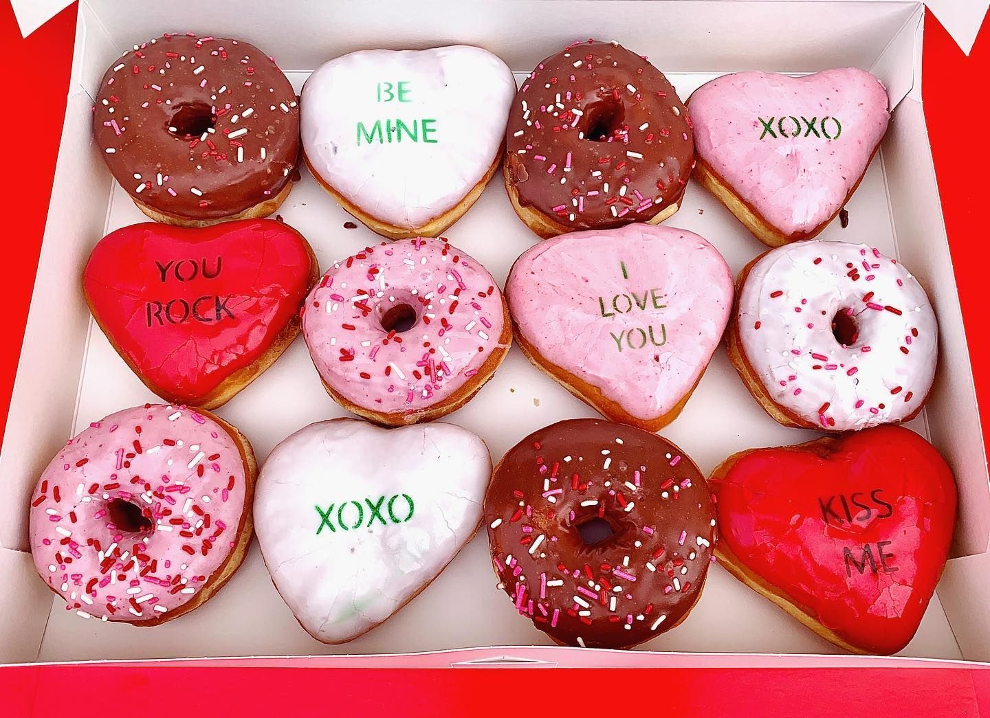 Box of Valentine's Day donuts: chocolate and pink frosted, heart-shaped with messages like "Be Mine" and "Kiss Me."