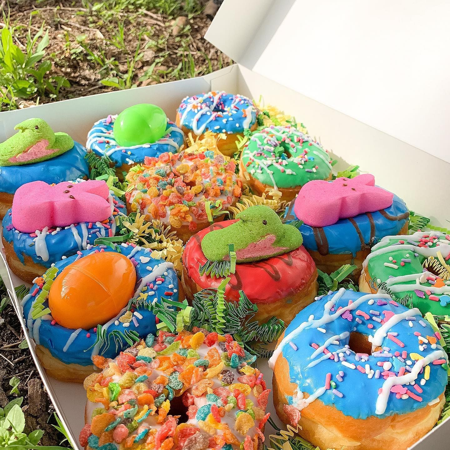 Box of colorful Easter donuts with various toppings, in an outdoor setting.