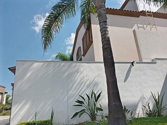 Pared blanca con sombras de palmeras, casa con ventana arqueada, cielo azul.