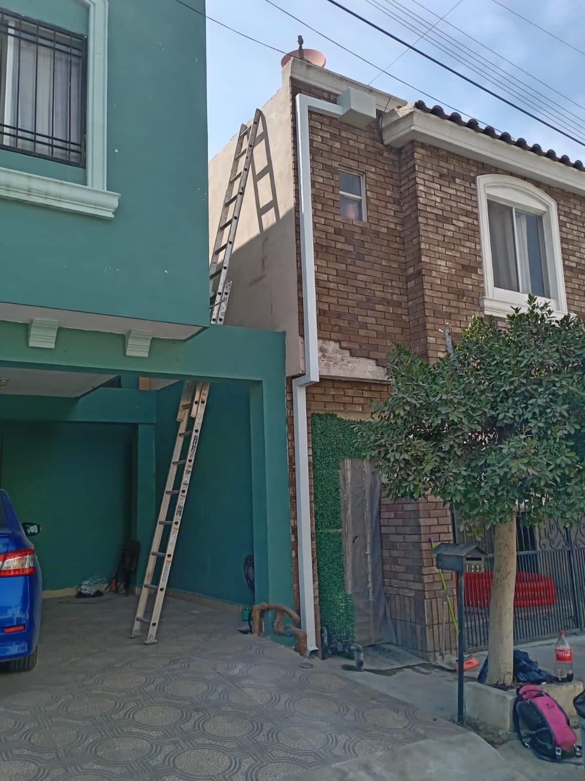 Edificios de ladrillo y vegetación de dos plantas con escaleras apoyadas contra ellos, un coche azul y un pequeño árbol delante.