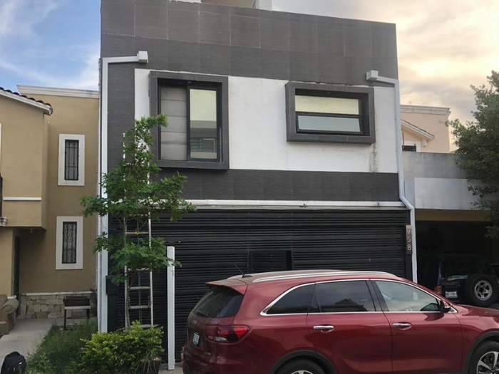 Casa de dos plantas con puerta de garaje negra, coche rojo aparcado delante, exterior gris y blanco.