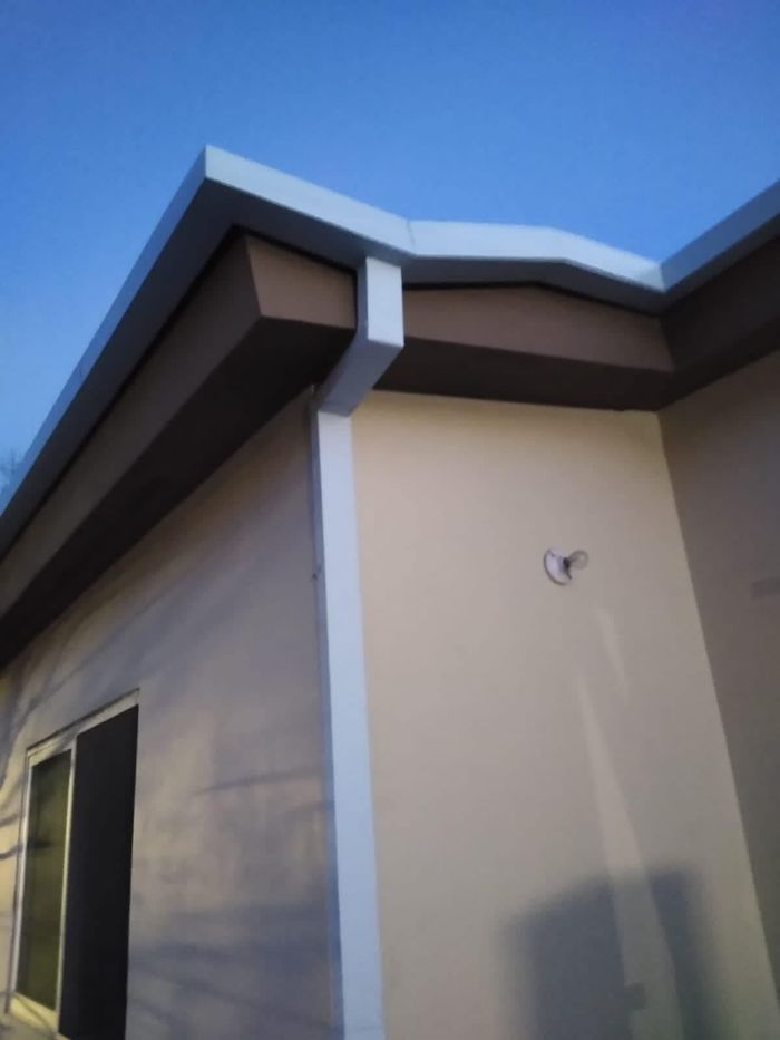 Esquina exterior de un edificio con alero marrón, canalones blancos y paredes beige contra un cielo azul.