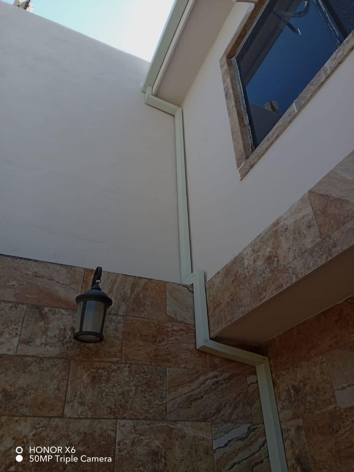 Esquina exterior de un edificio con canalones de color claro y una lámpara de pared sobre piedra beige y paredes blancas.