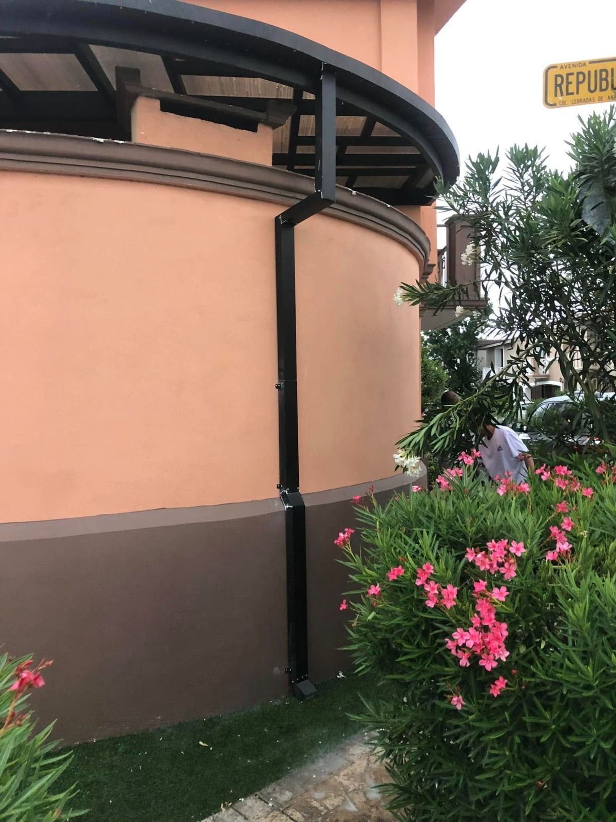 Vista exterior de una pared curva de color melocotón con una bajante negra, junto a vegetación y una señal de calle.