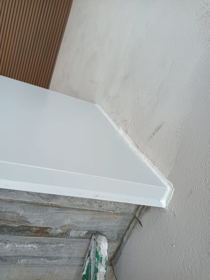 Estantería blanca con borde en ángulo contra una pared y un panel de madera. La estantería descansa sobre un soporte estructural.