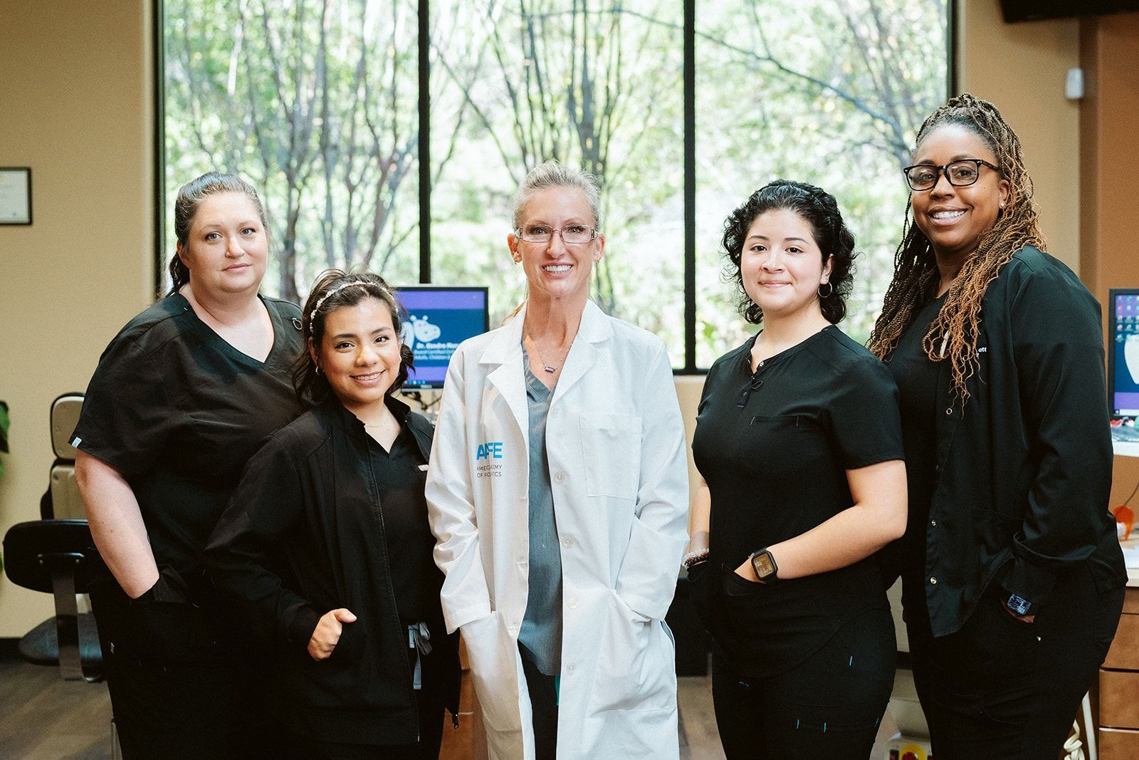 A group of five smiling staff members in medical scrubs and a white lab coat stand together in a bright office.