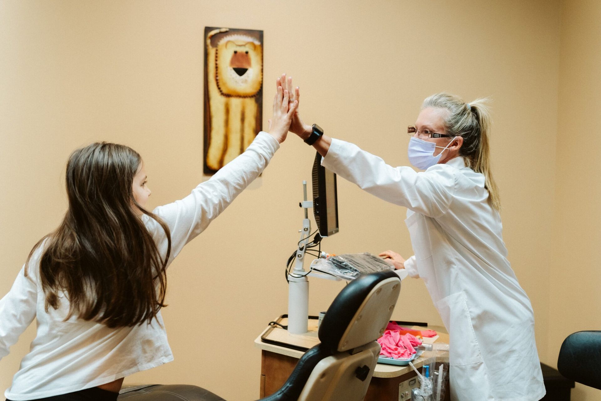 Girl and dentist in lab coat high-fiving in dental office; smiling, beige walls.