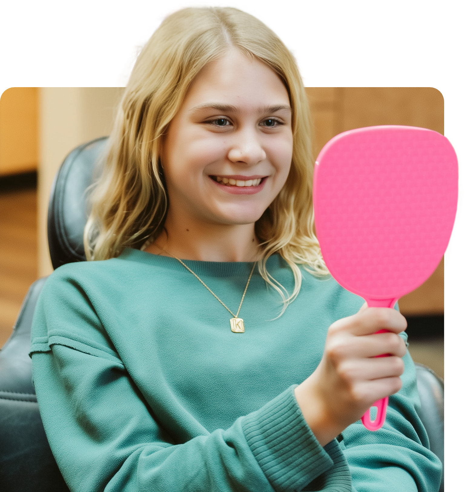 A smiling person in a teal sweater looks at their teeth in a pink hand mirror. They are in a dental office setting.