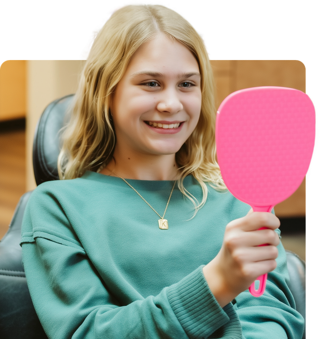 A smiling person in a teal sweater looks at their teeth in a pink hand mirror. They are in a dental office setting.